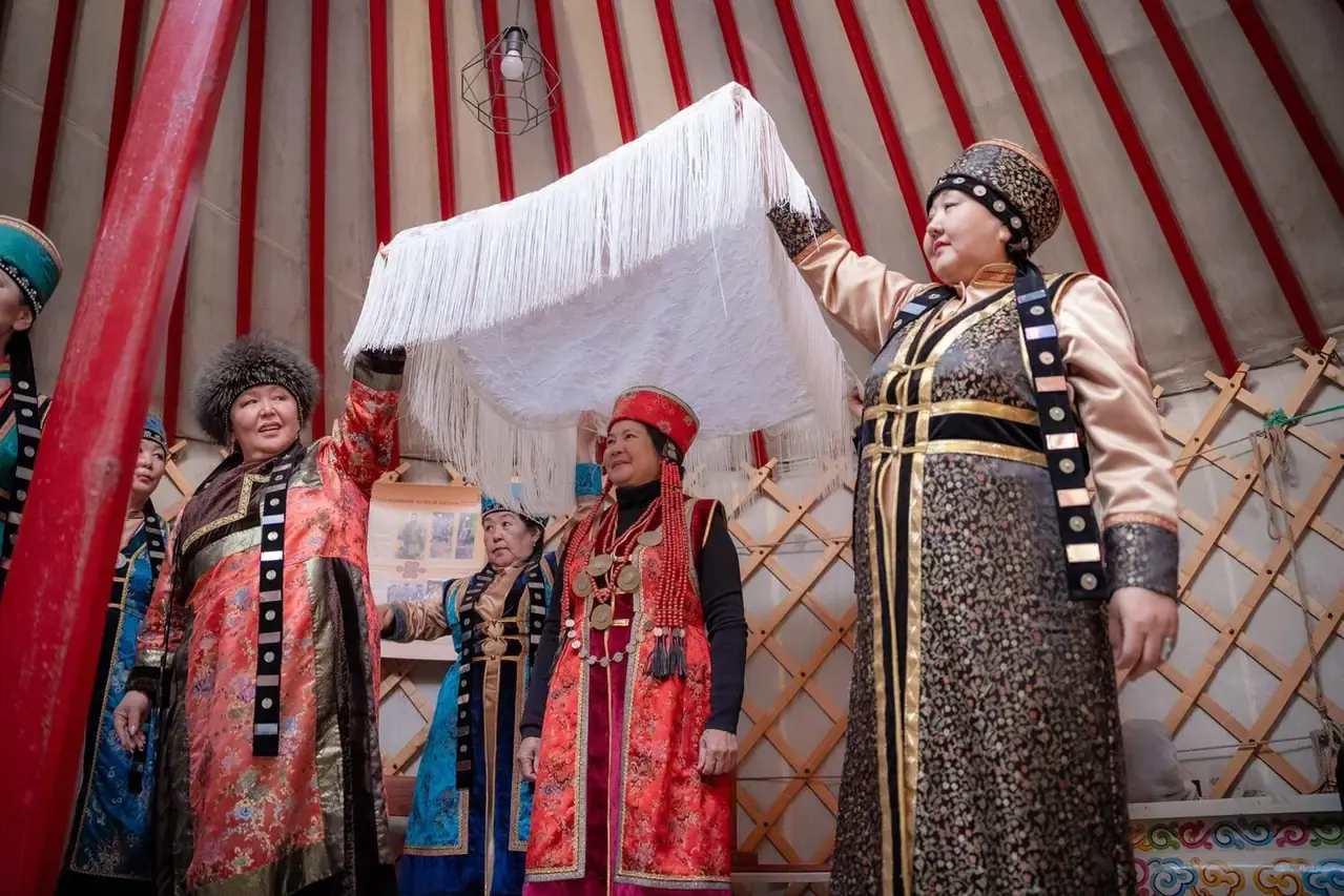 Phụ nữ Buryat trong trang phục truyền thống nhiều màu bên trong lều yurt, một người nâng tấm màn trắng